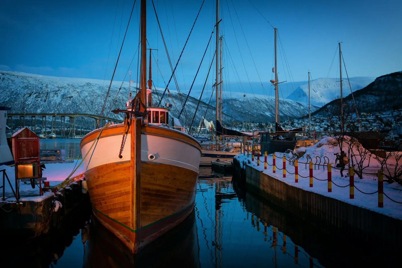 Wooden boat in Tromsø harbour with the bridge and snowy mountains at blue hour