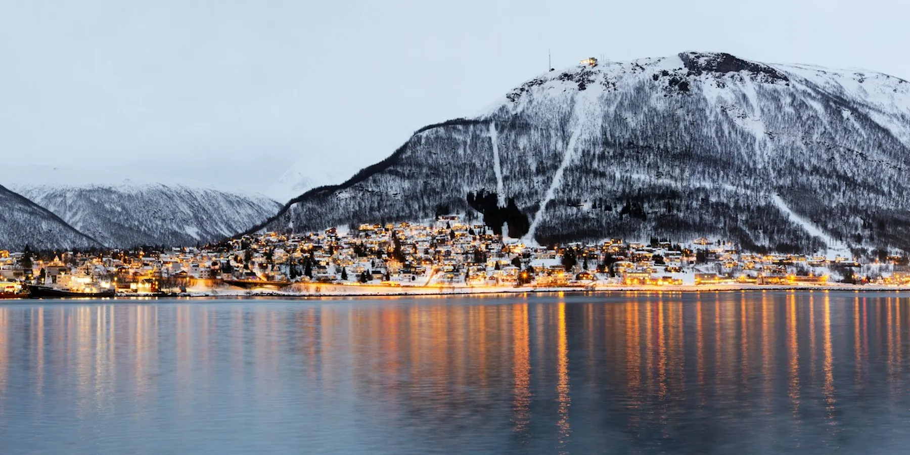 The harbour of Tromsø at dusk with city lights below a snow-covered mountain