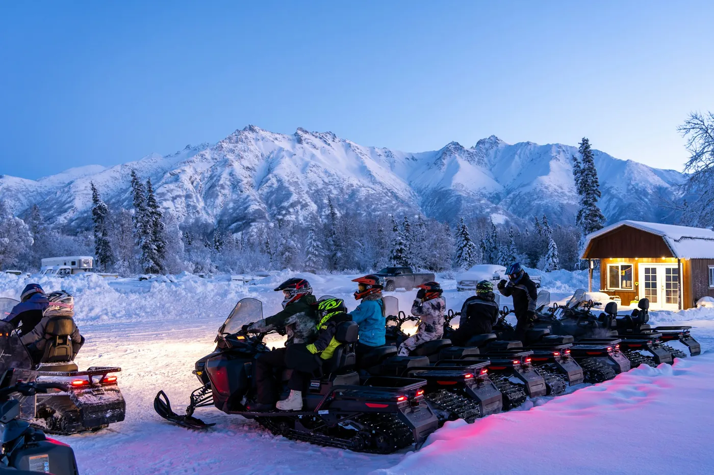 A group on snowmobiles in front of a wooden hut with mountains behind, in blue light